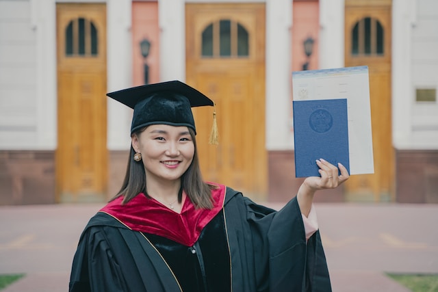 Eine Frau in Abschlusskappe und Talar hält ein Diplom in der Hand.