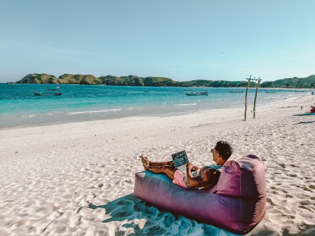 Eine Person sitzt auf einem lilafarbenen Sitzsack am Strand und benutzt ihren Laptop.