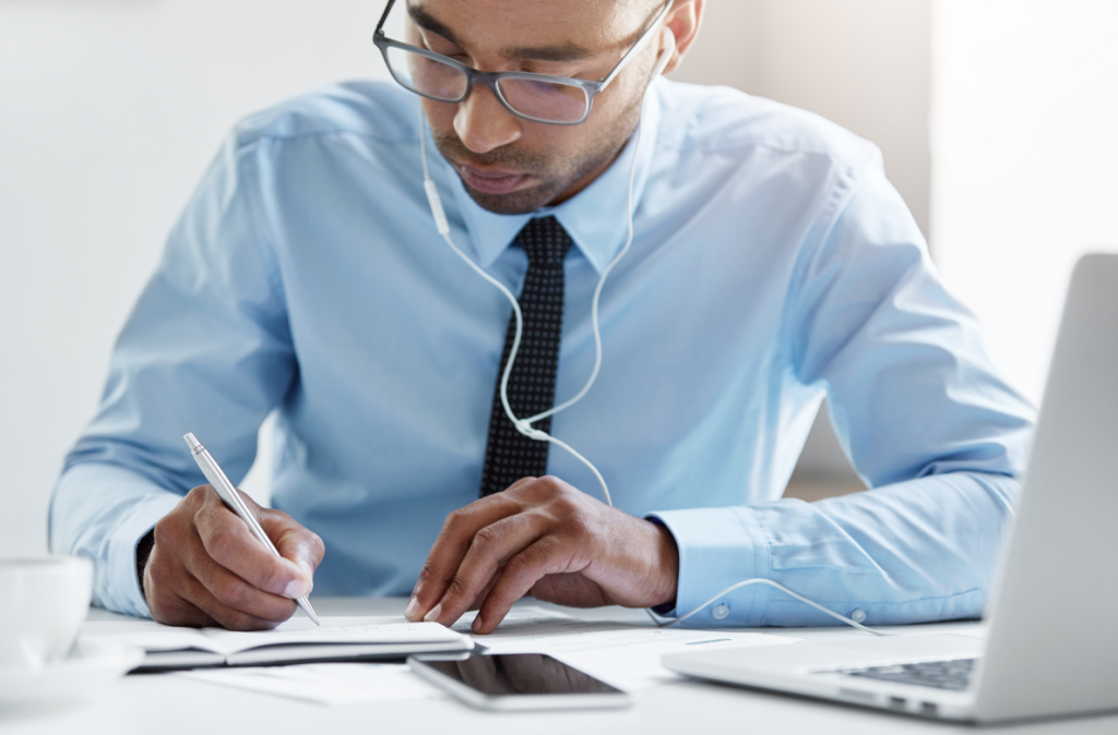 A man reviewing papers at a desk with a laptop, phone, and earphones.