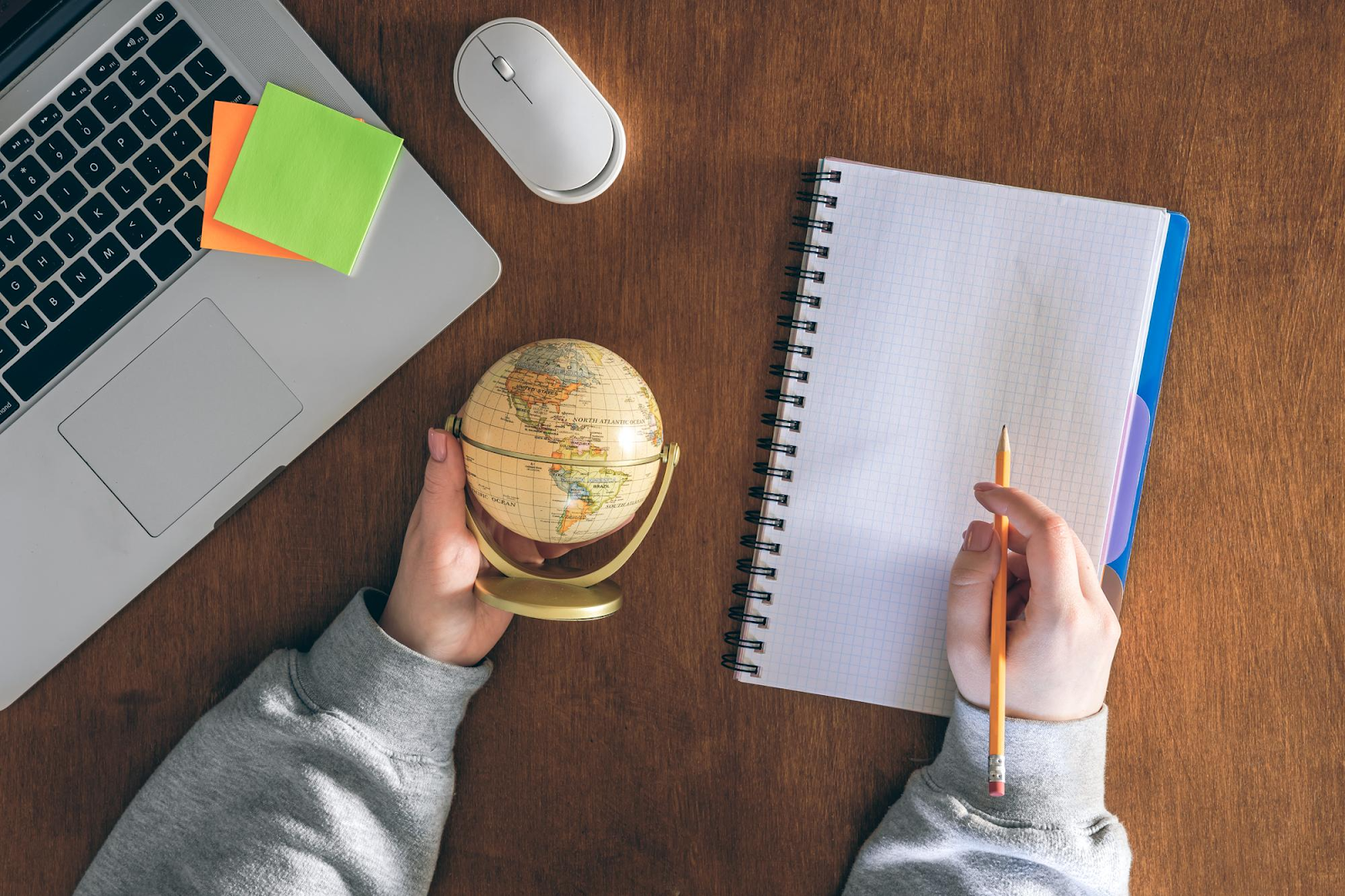 Une personne tient un globe terrestre dans une main et un crayon dans l'autre, assise à une table à côté d'une souris et d'un ordinateur portable.