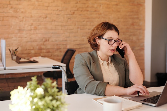 Donna con blazer e occhiali che parla al telefono mentre utilizza un computer portatile per tradurre un certificato di nascita.