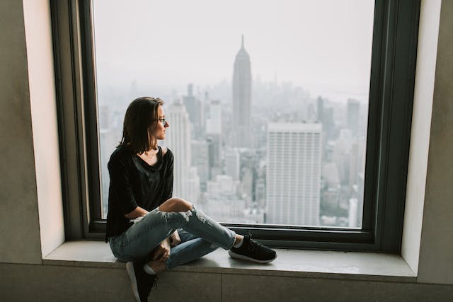 Donna con giacca nera e jeans blu denim seduta su una finestra di New York con vista.