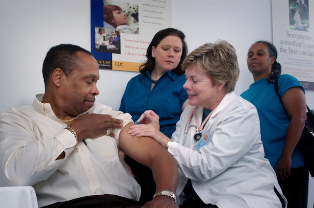 A man in a white shirt gets an injection on their arm from a doctor next to two onlookers.