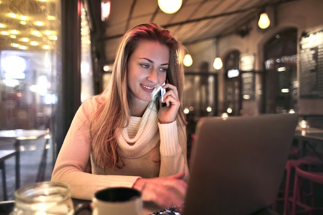 A relaxed woman in a cafe talking on a smartphone and using a laptop to look up translation services.