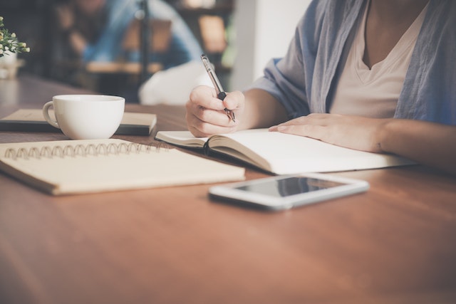 A woman writing on a notebook beside a teacup and a tablet computer.