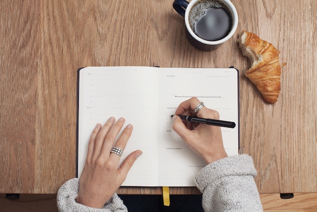 A picture of someone writing in a book on a table with a cookie and a cup beside it.