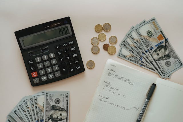 A photograph of a black calculator beside a book, pen, some coins, and dollar notes.