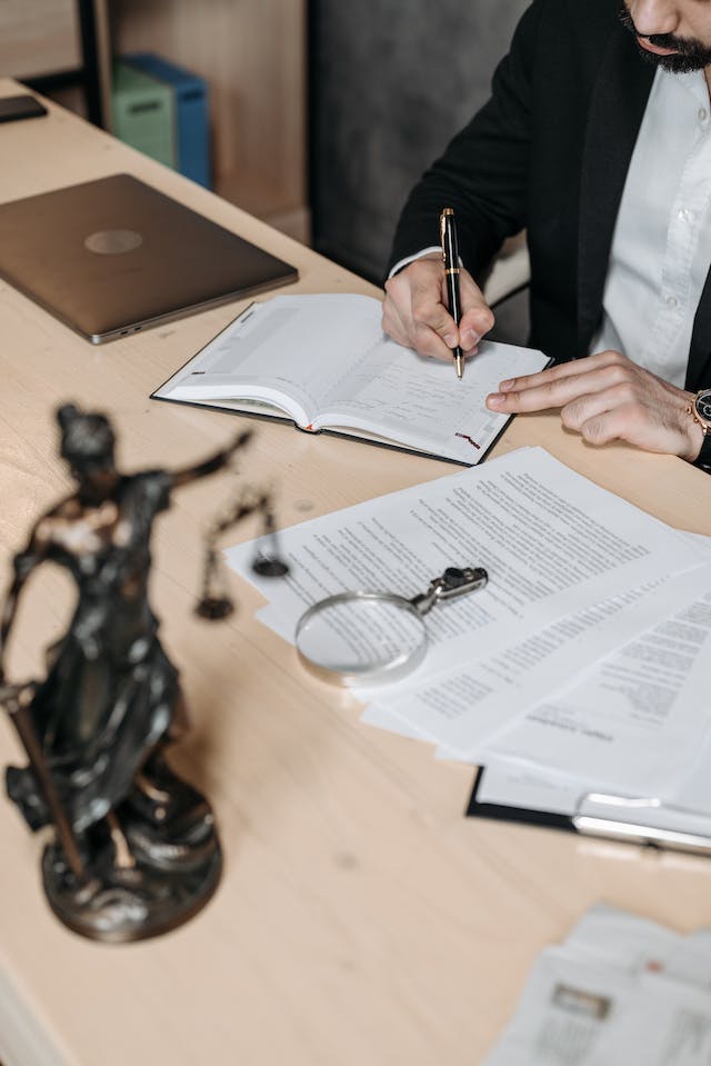 A man in a suit sitting at a table writing in a notebook while reviewing legal documents for translation.