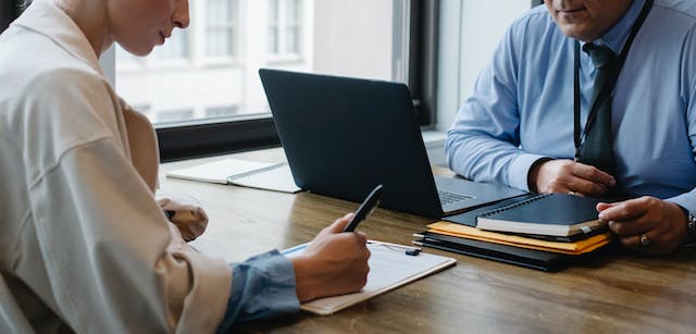 A picture of someone signing a document while another person looks on.