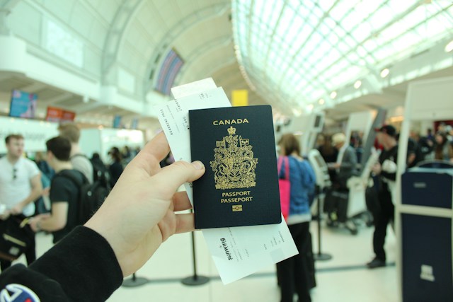 A person stands in a busy airport and holds out their Canadian passport with a boarding pass in it.