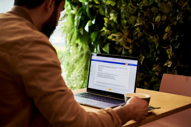 A man in a brown jacket holding a teacup while looking at texts on a laptop’s screen.
