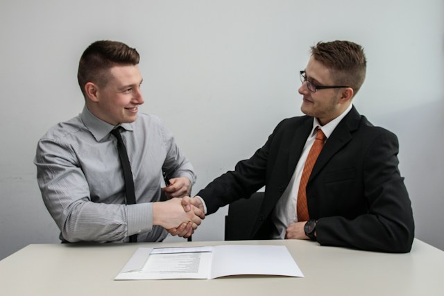 Two men face each other as they shake hands in agreement over a table that has a document.
