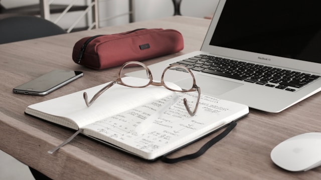 An opened laptop and notepad with glasses placed on a wooden table.