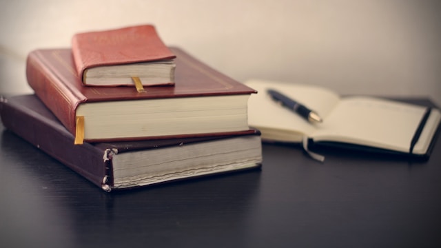 A stack of three books placed on a table beside an open book with a pen.