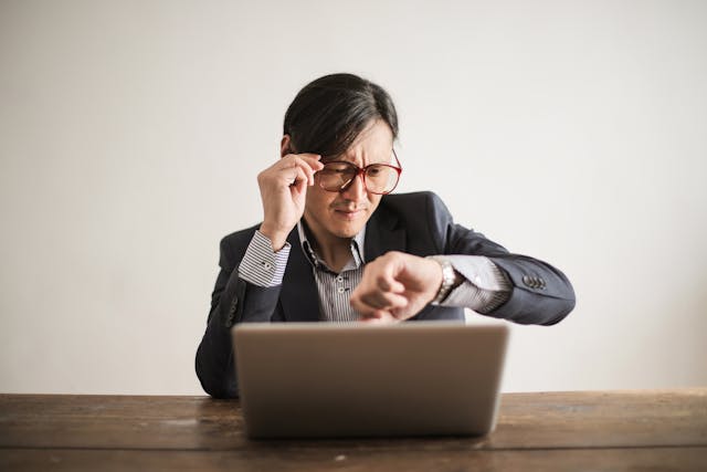 A man in a suit and glasses checks the time on his wristwatch.
