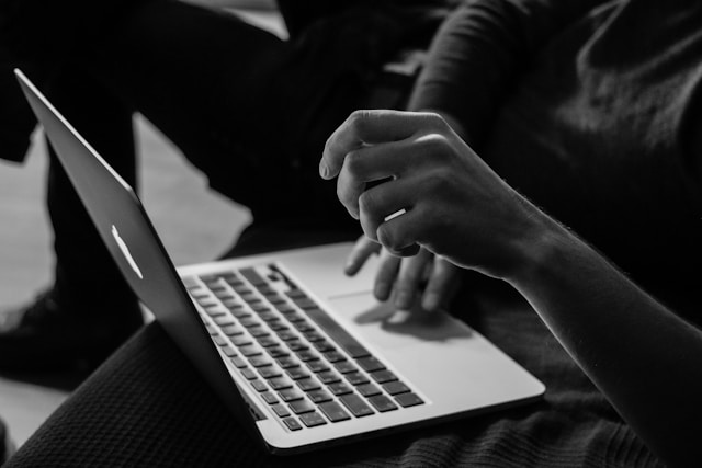 A person uses a MacBook while others linger in the background.
