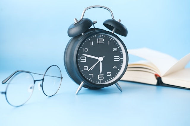 A black alarm clock with a pair of glasses and a book beside it.
