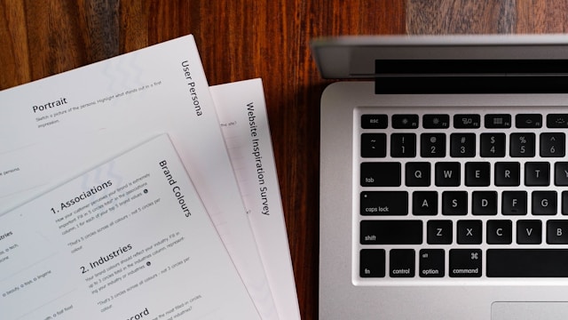 A pile of printed papers is beside a silver laptop on a wooden surface.
