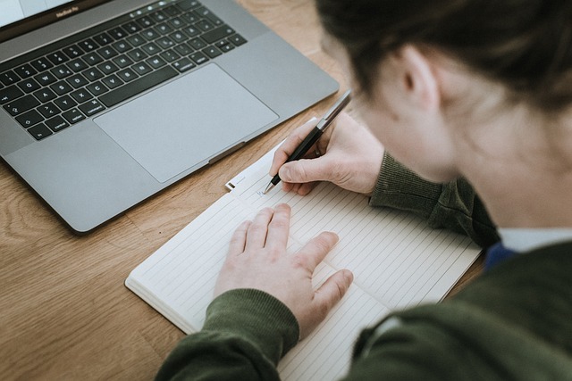 A person writes in a book before a silver laptop.
