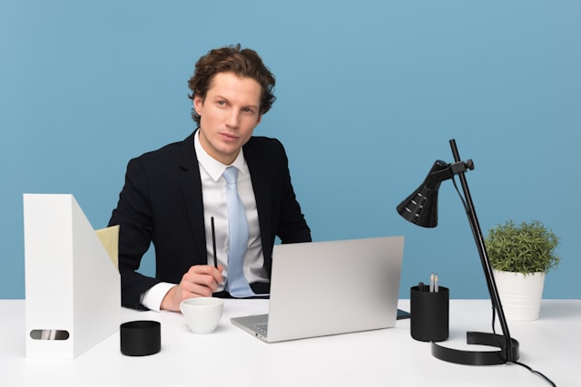 A person sits on a desk that has a laptop, a teacup, a vase, and other office items.