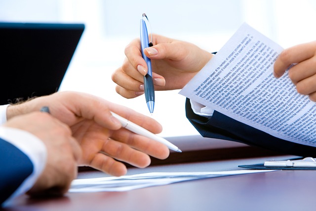 Two hands hold pens and sheets of printed paper opposite each other on a desk.
