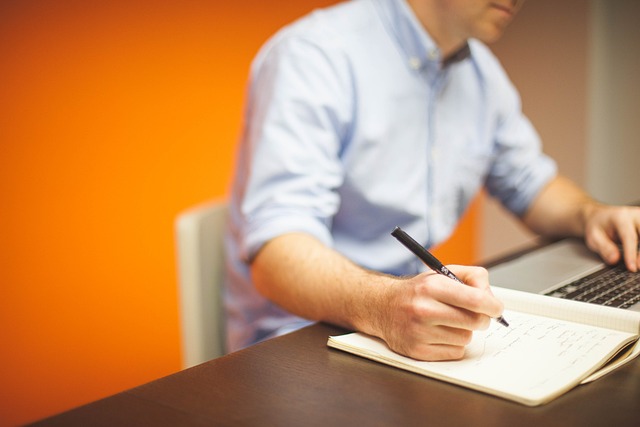 A person in a light blue shirt writes in a book beside a silver laptop.
