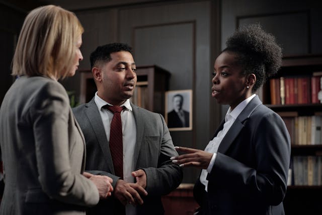 Three businesspeople in suits have a discussion while standing in an office.
