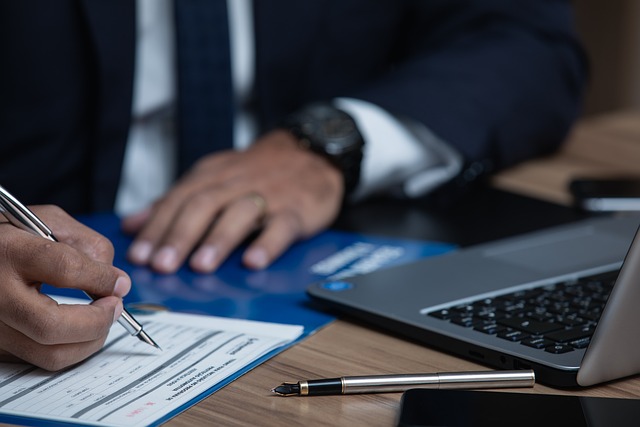 A person in a suit signs a document on a desk with a laptop in front of them.
