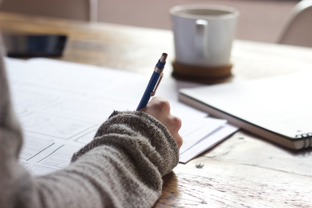 A person writes on white paper on a wooden table with a coffee cup on the table.
