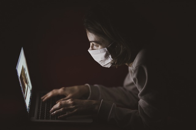 A person wearing a nose mask uses a laptop in a dark environment.
