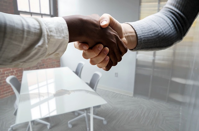 Two people shake hands with white chairs surrounding a table in the background.