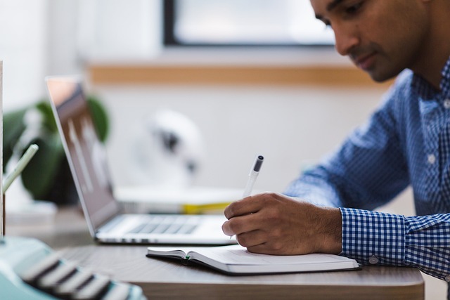 A person writes in a book beside a laptop.
