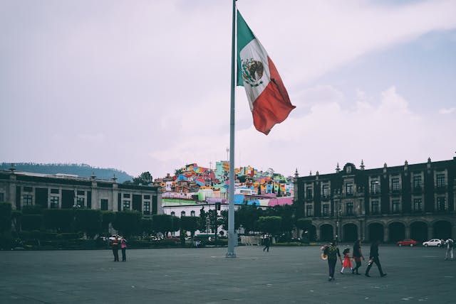 A huge Mexican flag flies in a square while people walk about.
