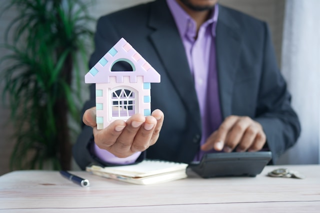 A person in a black suit holds a purple-colored house and uses a black-colored calculator.