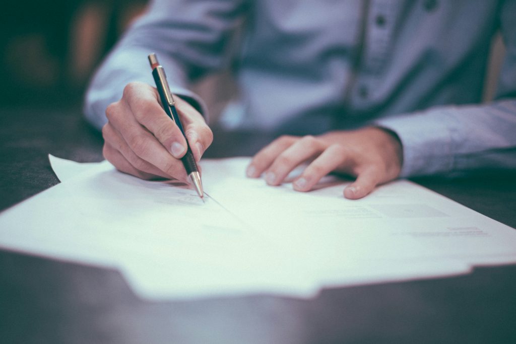 Man signing a document with a pen on a desk, close-up of hands filling out paperwork on white paper.
