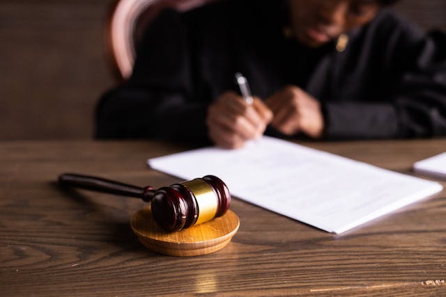 Безликий человек в судейской мантии подписывает документ на столе с помощью молотка.